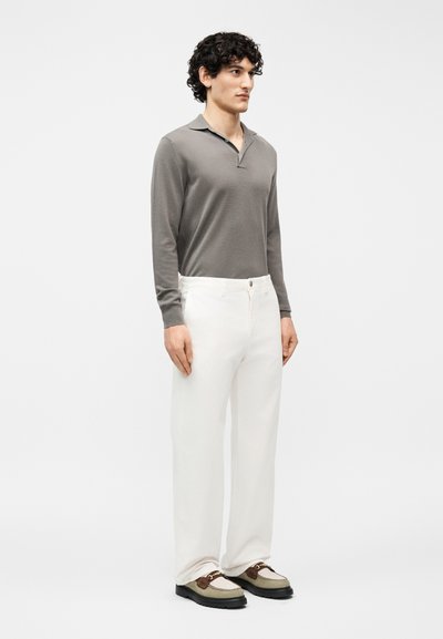 Homme aux cheveux bouclés portant un polo gris à manches longues, un pantalon blanc et des mocassins bicolores, debout devant un fond uni.