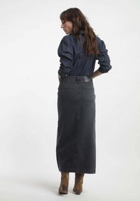 Jupe en denim noire longue avec poches arrière, taille haute et décoloration subtile. Associée à une chemise bleu foncé et des bottes de cowboy marron.