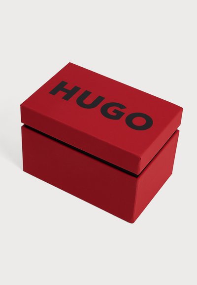 Boîte rectangulaire rouge avec couvercle noir, texte "HUGO" affiché sur un fond clair uni.