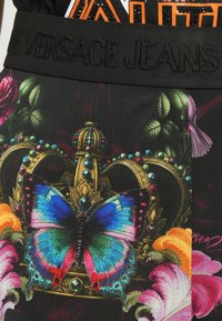 Čierna tkanina s farebnou motýlicou, zdobenou korunou a kvetinovými vzormi. Čierny pletený pás s embosovaným textom "Versace Jeans".