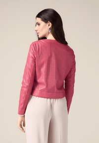 Giacca in pelle perforata rosa con colletto rotondo, polsini solidi rosa a contrasto e design aderente. Abbinata a pantaloni nei colori neutri.