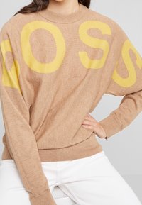 Hellbrauner Strickpullover mit großen gelben Buchstaben auf der Vorderseite, rundem Ausschnitt und gerippten Bündchen. Weicher Stoff, lässige Passform.