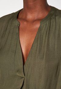 Blusa verde oliva con scollatura a V, in tessuto morbido e texture, con leggeri arricciamenti nel colletto, che evidenzia un aspetto semplice ed elegante.