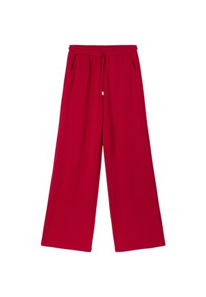 Jeune femme portant une doudoune blanche sur un crop top rouge et un pantalon rouge à jambes larges, debout devant un fond gris.