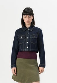 Giacca di jeans blu scuro cropped, con bottoni argentati, due tasche sul petto e cuciture visibili. Indossata sopra un top testurizzato bordeaux e una gonna oliva.