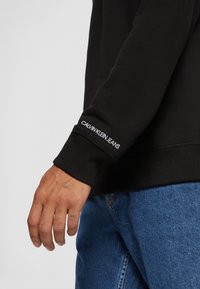 Bras gauche en sweat-shirt noir avec le texte « Calvin Klein Jeans » sur le bord de la manche, associé à un jean en denim bleu et un tatouage visible sur le doigt.