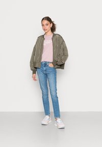 Casaco bomber verde-oliva, t-shirt rosa claro com texto, jeans skinny azuis e sapatilhas multicoloridas com solas brancas. Texturas suaves.