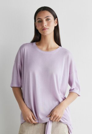 Frau mit langen dunklen Haaren, die ein hellviolettes Kurzarmoberteil trägt, das an der Taille gebunden ist, und vor einer schlichten weißen Wand steht.