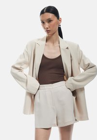 Blazer beige surdimensionné avec des manches retrouss�ées, associé à un débardeur marron côtelé et à un short beige taille haute. Design lisse et minimaliste.