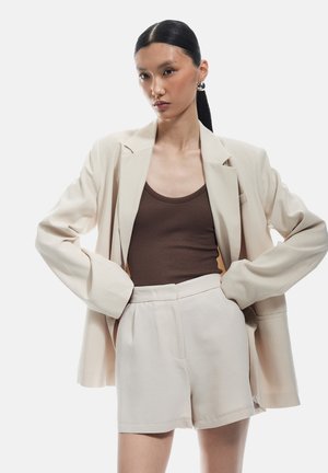 Blazer beige oversize con mangas enrolladas, combinado con una camiseta de tirantes marrón de canalé y pantalones cortos beige de cintura alta. Diseño suave y minimalista.