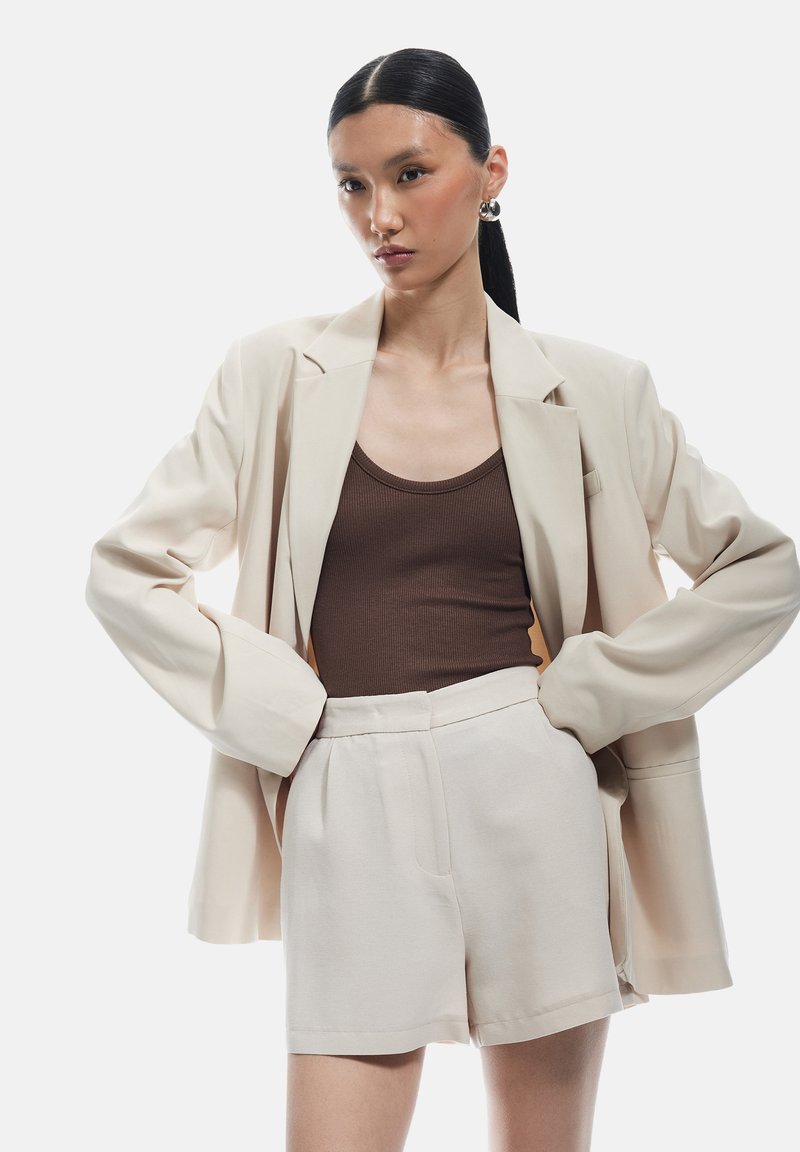 Blazer beige oversize con maniche arrotolate, abbinato a una canotta marrone a coste e pantaloni corti beige a vita alta. Design liscio e minimalista.