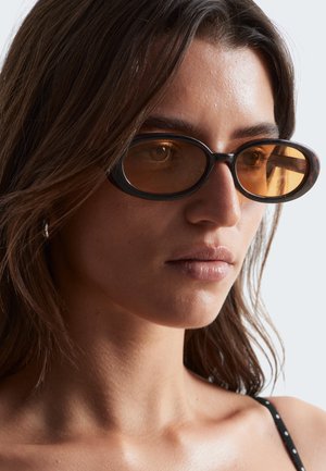 Jonge vrouw met lang bruin haar, een ovalen zwarte zonnebril met amberkleurige glazen en een zwart polkadot topje met bandjes, kijkt naar rechts.