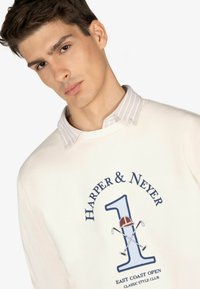 Sudadera crema con cuello redondo, que presenta un texto gráfico azul "HARPER & NEYER 1" y acentos relacionados con el golf. Superpuesta con una camiseta de rayas ligera.