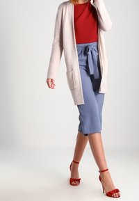 Lichtbeige gebreide cardigan met open voorkant, rode getailleerde top, hooggesneden blauwe broek met strik in de taille, en rode sandalen met hakken.