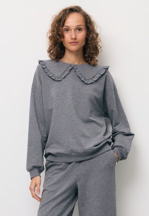 Kobieta z kręconymi włosami, ubrana w szarą bluzę z dużym falbankowym kołnierzem i pasujące szare spodnie, stojąca z jedną ręką w kieszeni.