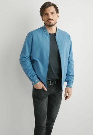 Mężczyzna z brodą w niebieskiej kurtce na czarnej koszulce i czarnych dżinsach, stojący z jedną ręką w kieszeni na jednolitym tle.