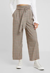 Pantalon taille haute à carreaux gris avec une ceinture à la taille, jambes larges et poches latérales. Porté avec des baskets montantes blanches.