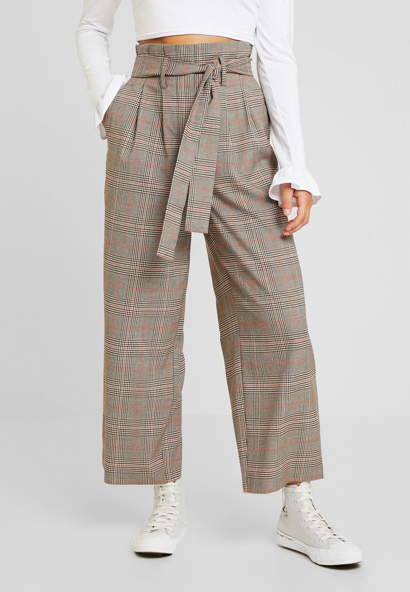 Pantalon taille haute à carreaux gris avec une ceinture à la taille, jambes larges et poches latérales. Porté avec des baskets montantes blanches.