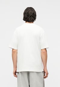 Άτομο με μεσαίου μήκους σκούρα μαλλιά, που φοράει απλό λευκό κοντομάνικο πουκάμισο και ανοιχτόγριζο παντελόνι, φαίνεται από πίσω.