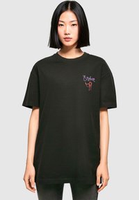 Camiseta negra de gran tamaño hecha de algodón, con "Ozzfest '97" bordado en púrpura y rojo en el lado izquierdo del pecho. Manga corta, cuello redondo.