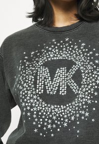 Sudadera gris con un diseño redondeado de estrellas plateadas que presenta un logo "MK" en el centro, hecho de lentejuelas y piedras brillantes.