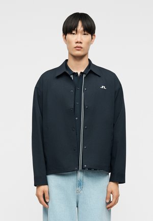 Mladý muž oblečený v čiernej zapinanej bunde cez čierne tričko s bielymi pruhmi a svetlomodrými džínsami, stojaci pred jednoduchým pozadím.