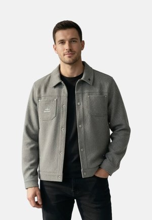 Mand iført en grå tekstureret jakke med knapper over en sort skjorte, stående med den ene hånd i lommen mod en ensfarvet baggrund.
