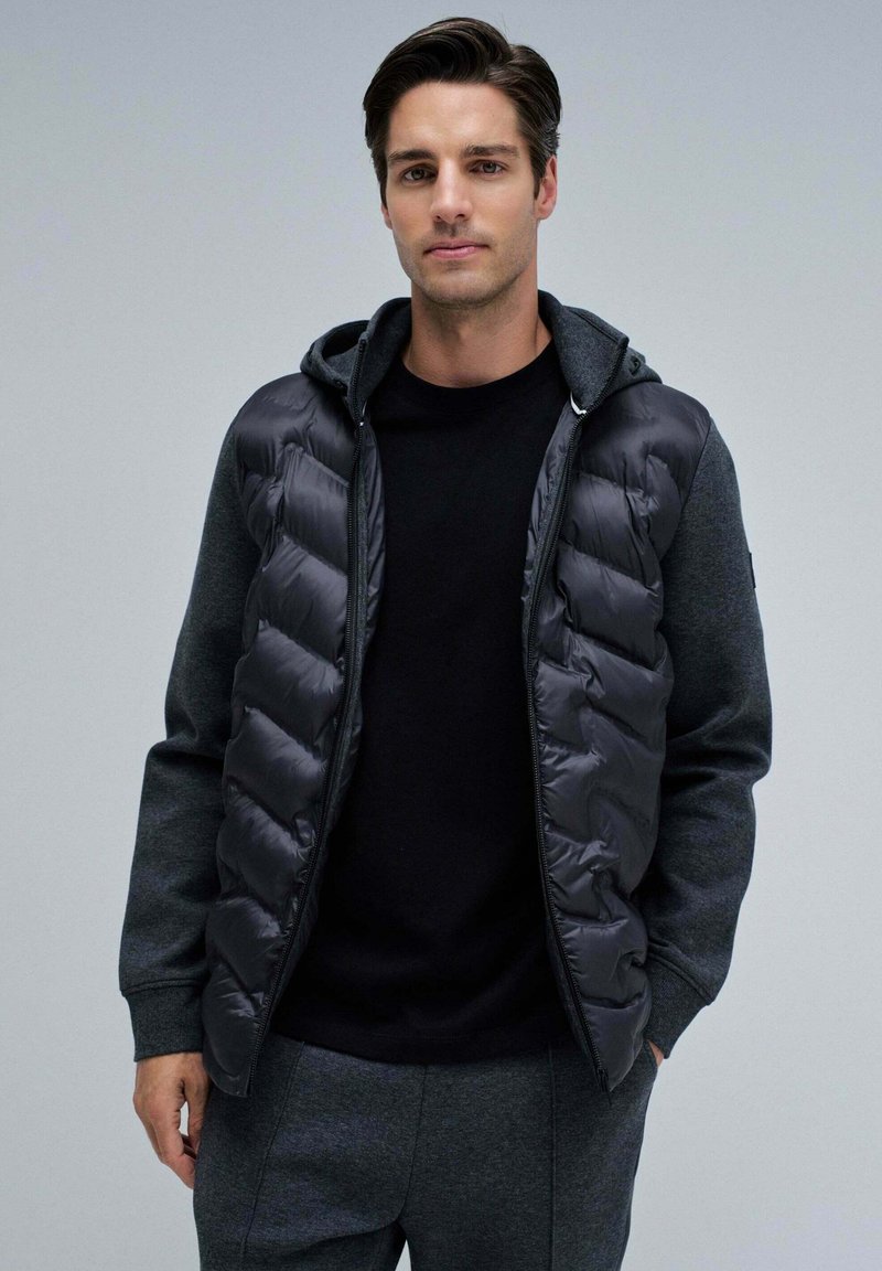 Gilet en duvet gris foncé avec un design matelassé et une capuche, superposé sur une chemise noire. Le modèle porte un pantalon gris assorti, créant un look monochromatique.