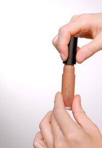 Tube de gloss à lèvres liquide avec un capuchon noir, tenu entre deux doigts. Le gloss a une couleur nude pastel avec une finition brillante et une forme cylindrique.