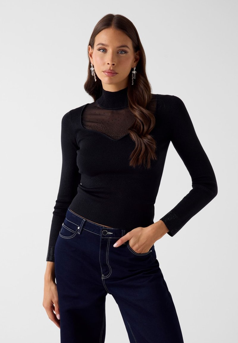 Junge Frau mit langem braunem Haar trägt ein schwarzes Langarmshirt mit durchsichtig verziertem Ausschnitt und dunkelblaue, hoch taillierte Hose.