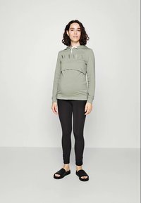 Sweatshirt de maternité en vert olive avec une poche kangourou et l'impression "MAMA", associé à des leggings noirs et des sandales noires. Texture de tissu lisse.