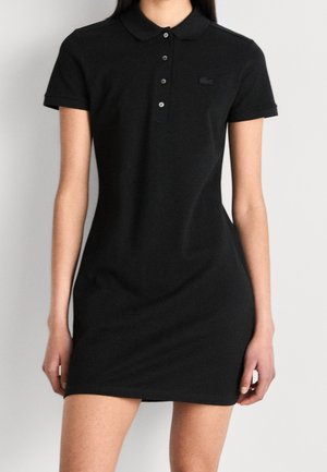 Frau in einem enganliegenden schwarzen Polokleid mit kurzen Ärmeln, Kragen und Knöpfen vorne.