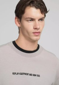 Felpa grigio chiaro con collo rotondo nero. Presenta testo ricamato "REPLAY-EQUIPMENT AND DNM GDS" sul petto. Texture liscia.