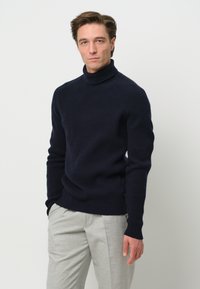 Dunkelblauer gerippter Rollkragenpullover aus einem strukturierten Wollgemisch. Kombiniert mit hellgrauen Hosen mit geradem Bein.