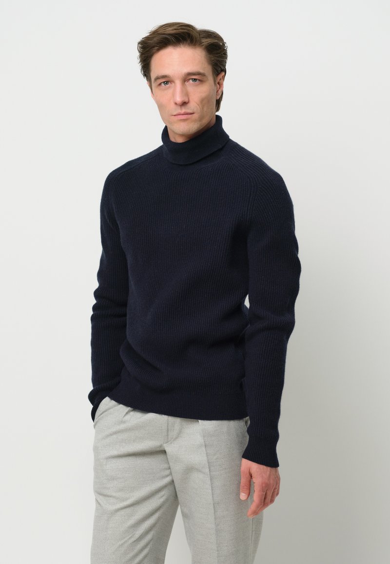 Dunkelblauer gerippter Rollkragenpullover aus einem strukturierten Wollgemisch. Kombiniert mit hellgrauen Hosen mit geradem Bein.