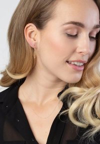 Gouden oorbellen in de vorm van veren hangen aan de oorlel van het model. Het verfijnde ontwerp contrasteert met haar zwarte shirt en zachte, golvende blonde haar.