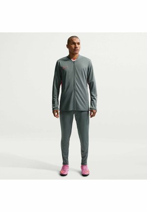 Homme portant un survêtement Nike gris avec veste zippée et pantalon assorti, debout, chaussures de sport roses, fond uni clair.
