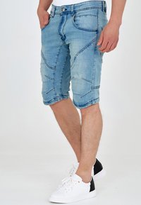 Pantaloni scurți din denim albastru deschis, cu un design ajustat, având multiple modele de cusături și o lungime până la genunchi. Asortați cu adidași albi și negri.
