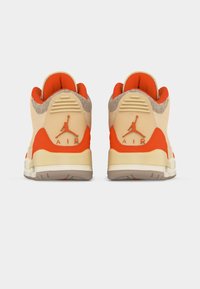 Ζευγάρι μπεζ και πορτοκαλί αθλητικά παπούτσια Air Jordan, εμφανιζόμενα από πίσω με το λογότυπο Jumpman προς τα πάνω και το κείμενο «AIR» στις πλευρές της φτέρνας.