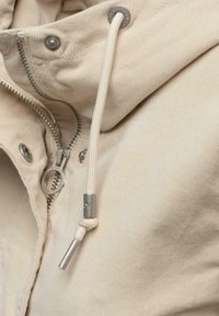 Beige Cordjacke aus Cord mit einem silbernen Reißverschluss und Druckknöpfen, ausgestattet mit einem weißen Kordelzug mit Metallenden und strukturiertem Stoff.