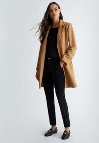 Cappotto cammello, maniche lunghe, chiusura con un singolo bottone; abbinato a pantaloni neri in velluto e mocassini neri con accenti dorati.