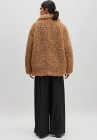 Flauschige, braune, übergroße Jacke mit hohem Kragen, die eine dicke, weiche Textur aufweist. Kombiniert mit lockeren schwarzen Hosen und klobigem Schuhwerk.