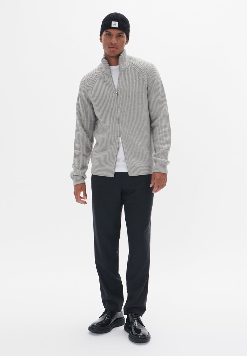 Grau gestreifter Zip-Up-Pullover mit hohem Kragen, kombiniert mit einem weißen Hemd und dunklen Hosen. Schwarze Schuhe vervollständigen das Outfit. Schwarze Mütze auf dem Kopf.