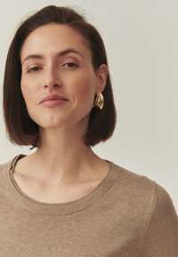 Pull en maille marron avec col rond. Le modèle porte une grande boucle d'oreille dorée brillante. Texture lisse et design minimaliste.