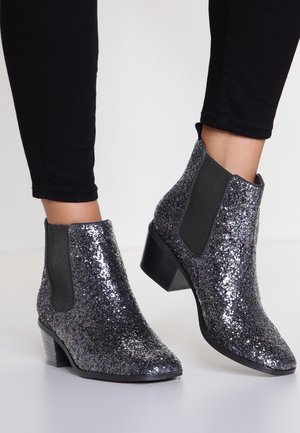 Bottines noires brillantes avec paillettes, panneaux latéraux élastiques et talons blocs bas, portées avec un pantalon noir ajusté.
