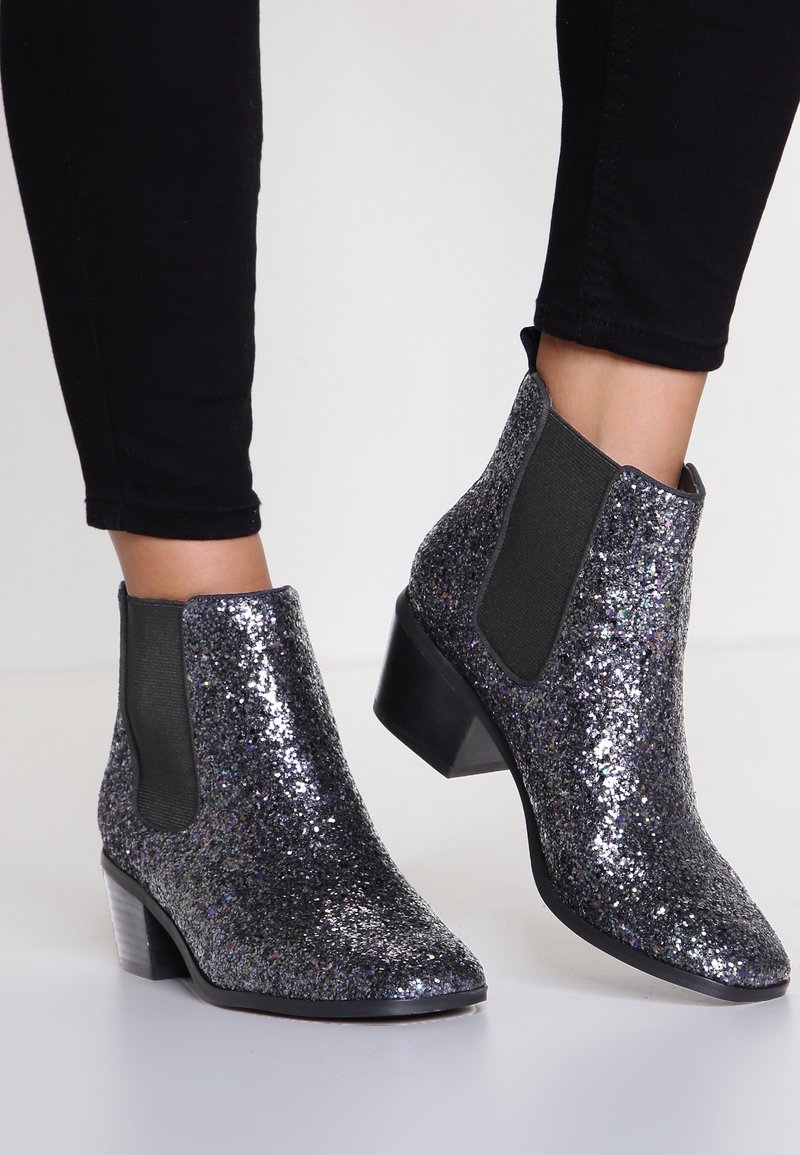 Bottines noires brillantes avec paillettes, panneaux latéraux élastiques et talons blocs bas, portées avec un pantalon noir ajusté.