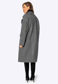 Lungo cappotto grigio in lana con un ampio colletto, vestibilità comoda e cuciture a vista, indossato sopra stivali neri alti fino alla coscia. Design minimalista, senza chiusure visibili.