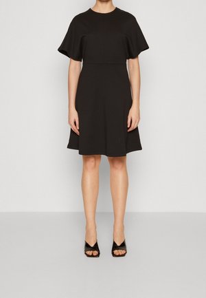 Mujer con vestido negro hasta la rodilla, con mangas cortas y anchas, y sandalias negras de tacón con punta abierta, de pie frente a un fondo liso.