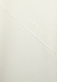 Tessuto crema con una superficie liscia, caratterizzato da una cucitura diagonale e lettere in rilievo "CPNHGN" dello stesso colore.