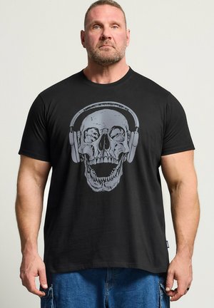 Mann mit kurzen Haaren und Bart, der ein schwarzes T-Shirt mit einem grauen Totenkopf mit Kopfhörern trägt, steht vor einem schlichten hellen Hintergrund.